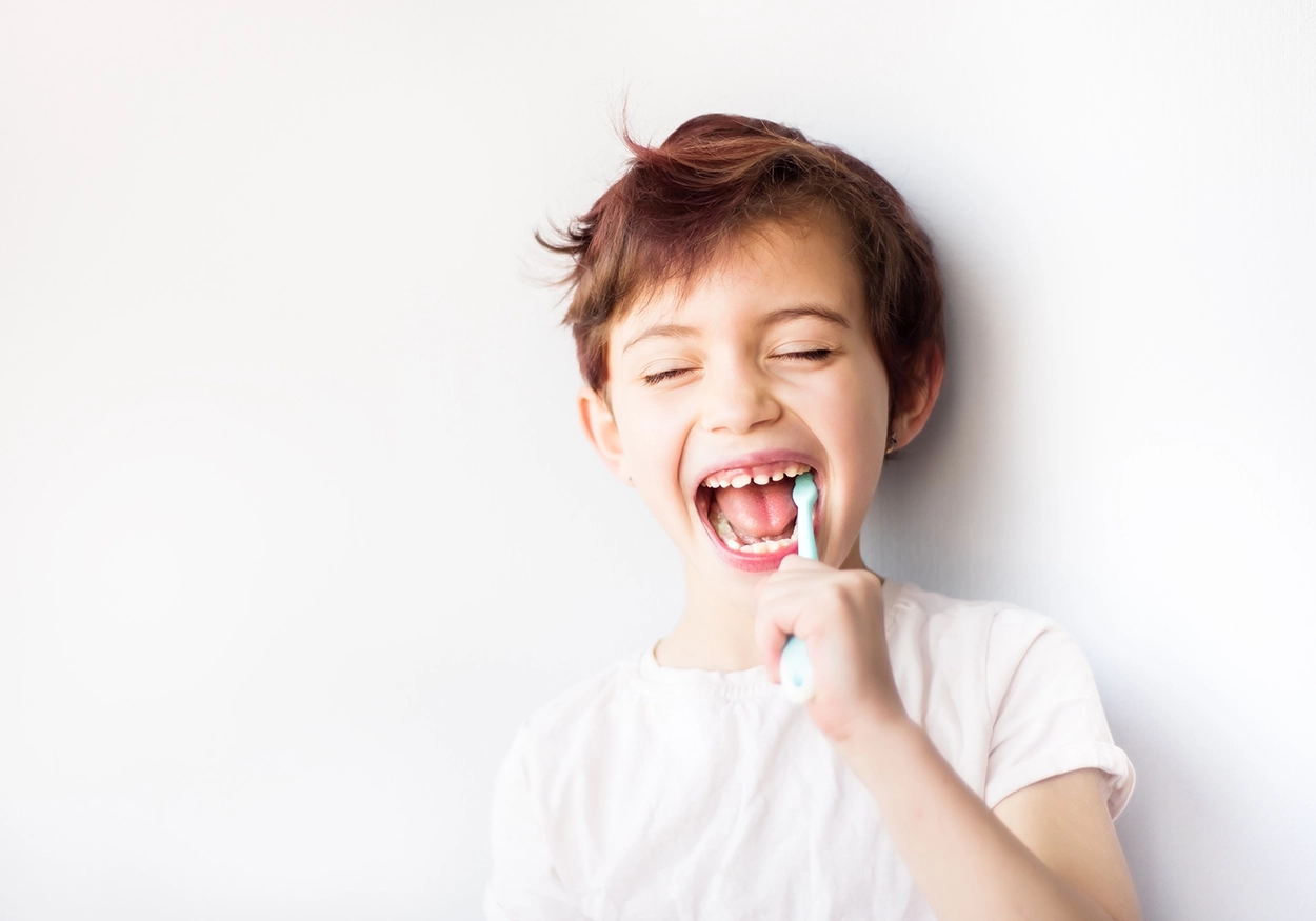Ein Bild von einem Kind mit lächelndem Gesicht und geschlossenen Augen, das sich die Zähne mit einer blau-weißen Zahnbürste putzt, ein T-Shirt trägt und sich vor einer Wand befindet.