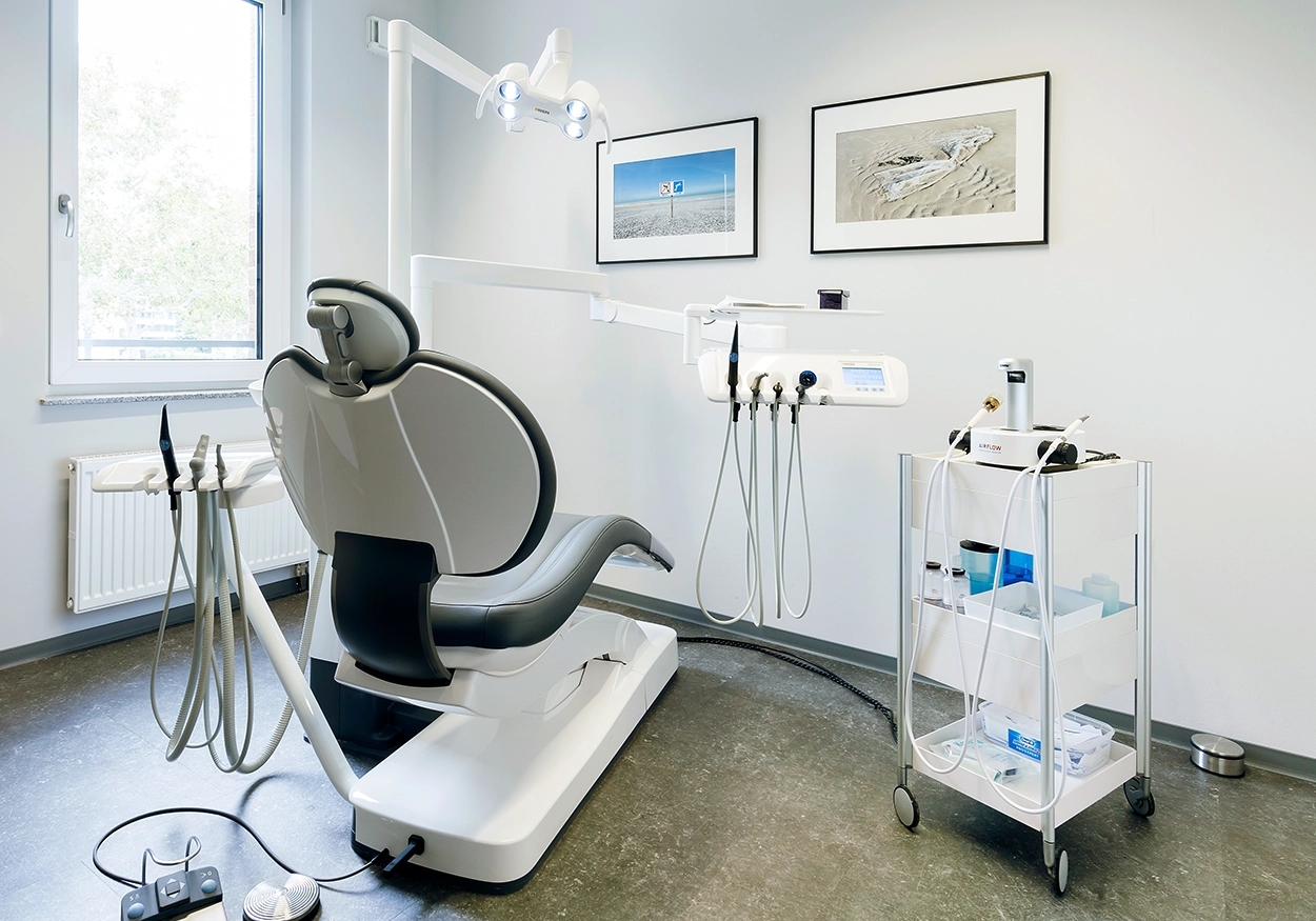 Ein Bild, das einen Behandlungsraum mit Behandlungsstuhl, zahnmedizinischen Instrumenten, eine Wand mit Bilder nund ein Fenster zeigt.